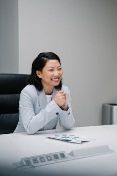 A smiling woman in business attire sits at a desk in an office environment.